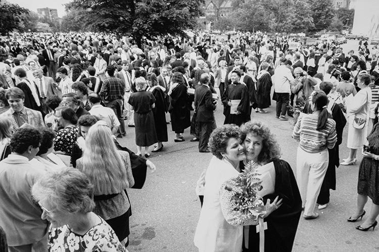 A large crowd of students and families, wearing 1990 fashions with permed hair, chat and hug on Front Campus.
