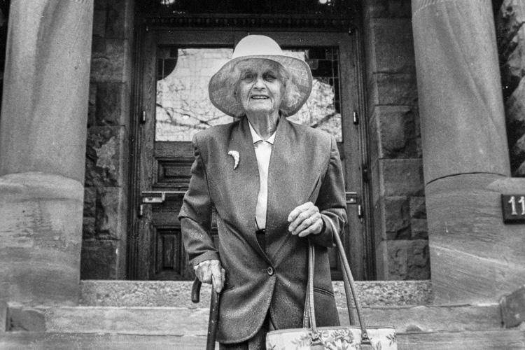 Clare Stephenson, holding her cane and wearing a big hat, smiles in front of the door to Woodsworth College.