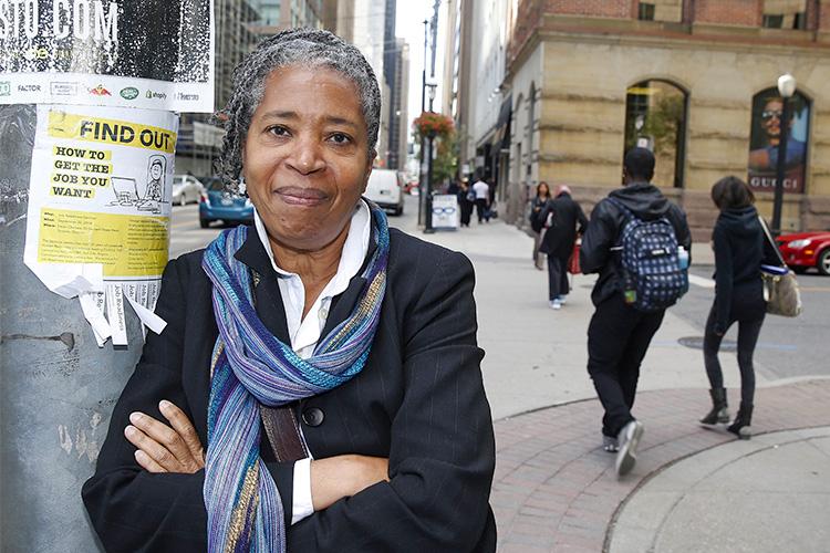 Dionne Brand smiles as she leans, arms folded, on a telephone pole on a downtown street. The sign on the pole reads: "How to get the job you want"