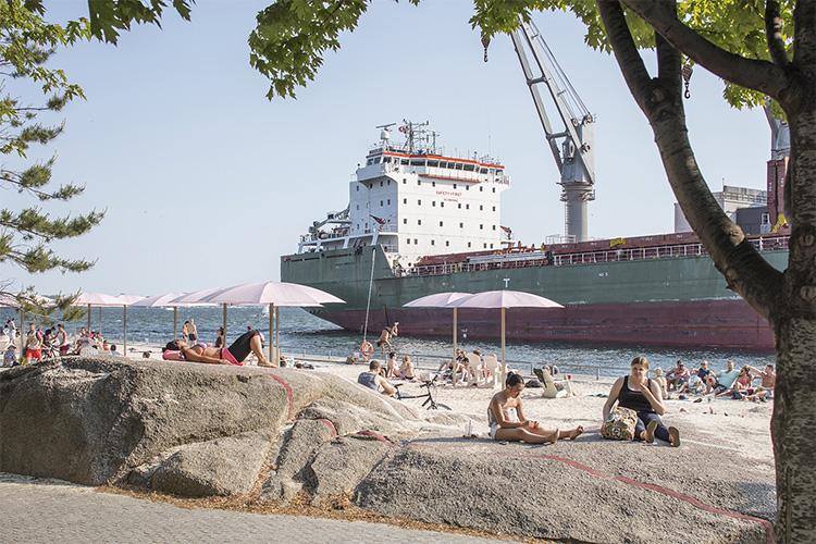 People relax on a sandy beach under umbrellas, right across from a huge cargo ship unloading.