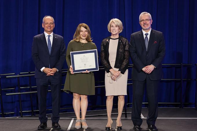 Barbara Dick stands on stage holding her Award of Excellence, next to President Gertler, Chancellor Patten and Scott MacKendrick (photo by Gustavo Toledo Photography)
