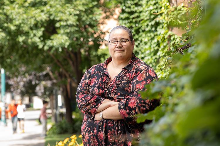 Susan Hill looks thoughtful while standing on a sidewalk with trees and bushes behind her.