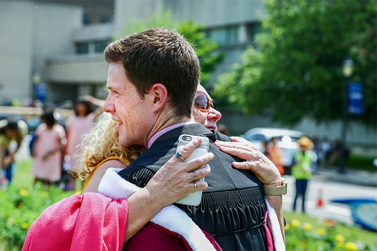 A young man in hood and gown smiles as a woman hugs him.