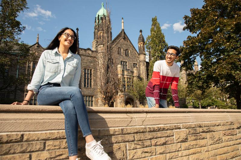Piya Sial and Shashwat Aggarwal laugh while sitting, socially distanced, on a stone wall outside Trinity College.