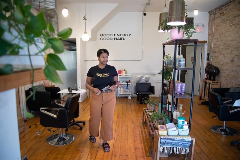 Allison Hill stands in a hair salon wearing a t-shirt that reads: restore. We see green plants and a sparkling wood floor.