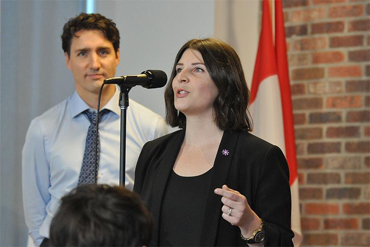 Justin Trudeau looks on as Melissa Sariffodeen speaks into a microphone.