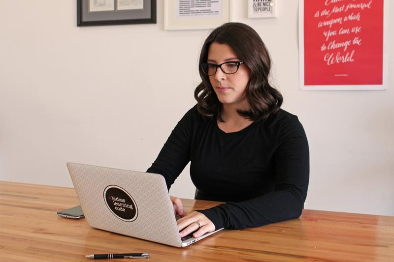 Melissa Sariffodeen types intently on a laptop. The laptop has a big sticker that reads Ladies Learning Code.