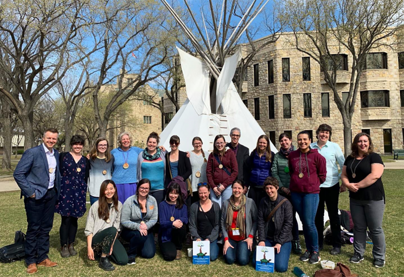 A group of people smile as they stand in front of a tipi on a lawn at U of T.