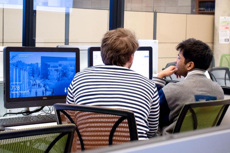 Two young men gaze thoughtfully at a computer screen in a study room full of tables and chairs