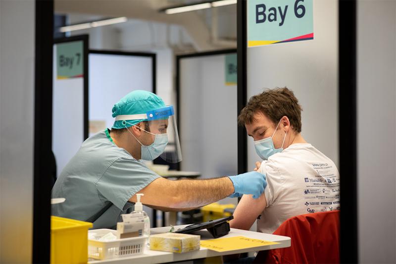 Stefan Juckes holds up his sleeve as a health-care worker in scrubs, cap, mask, face shield and gloves administers a needle.