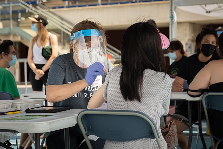 A young woman receives a vaccination in her arm from professor Lynn Wilson, wearing mask, face shield and gloves.