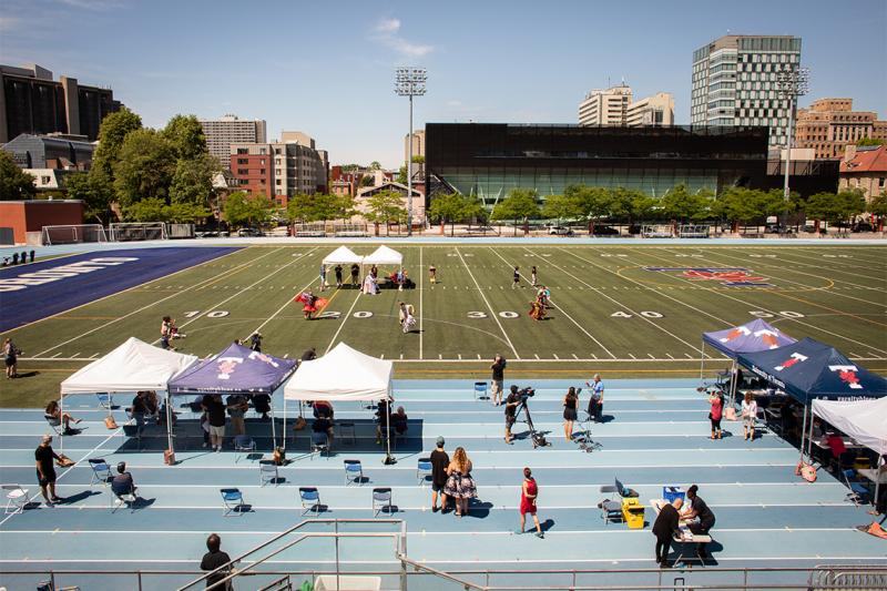 Dancers whirl across the Varsity Stadium football field while people get vaccinated at tables set up under canvas shelters.