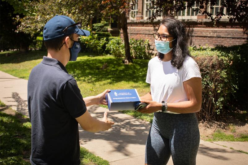 On a sidewalk, Brian Desrosiers-Tam hands a box with the U of T logo on the top to Devni Kumarasinghe. Both wear masks.