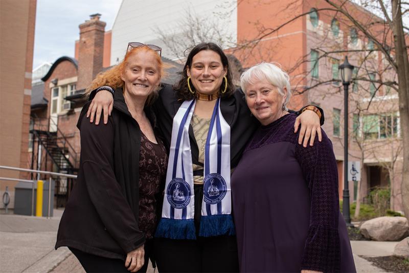 Caeley Genereux smiles happily, wearing her graduation scarf and with arms around two older women.