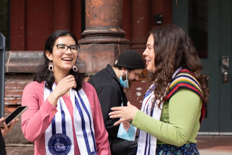Kristine Keon and Angelique Belcourt stand together, laughing. They wear U of T Indigenous grad scarves.