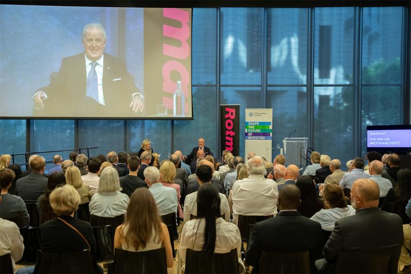 An audience watches as Brian Mulroney speaks on a stage before a large window.