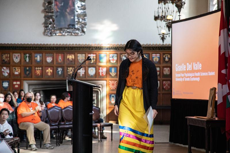Giselle Del Valle, wearing an orange shirt and a traditional ribbon skirt, steps up to the podium.