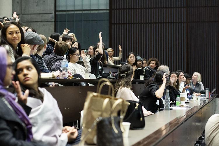 Women sitting in a tiered lecture theatre raise their hands to be heard, smiling at each other.