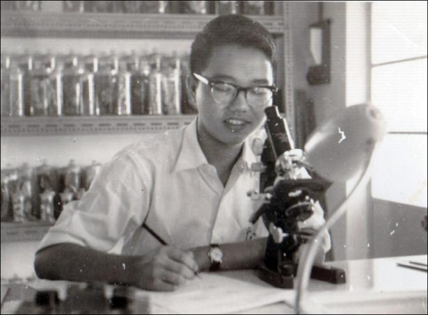 A young C.C. Liew smiles as he writes on a notepad while peering into a microscope, sitting in front of shelves filled with sample bottles.