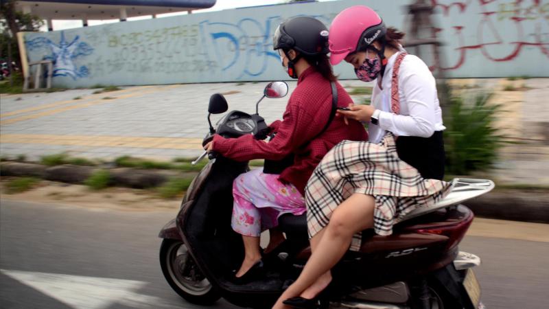 Two women in skirts and helmets ride a motorcycle. The woman in the rear is looking down at her smartphone.