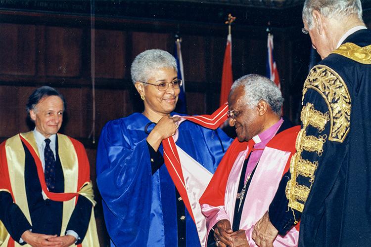 Desmond Tutu smiles as Mary Anne Chambers lifts an academic hood over his head. Dignitaries in robes look on.
