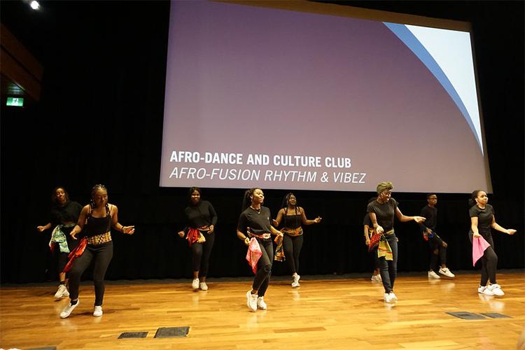 8 people dance on a stage beneath a banner that says: Afro-Dance and Culture Club, Afro-fusion rhythm and vibez.