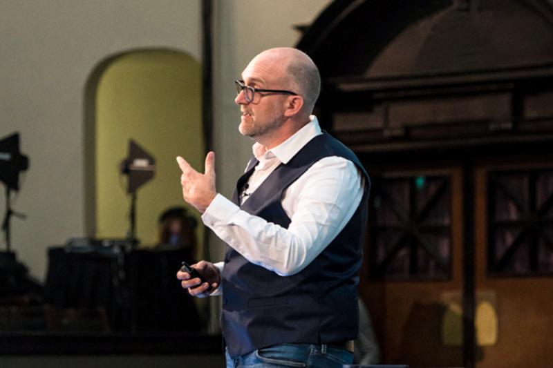 A man gestures while speaking at the front of a room.