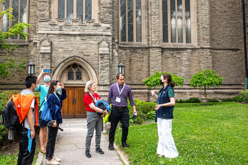 A group of people chat with a tour guide while one man takes a photo, on a lawn outside an old stone building.