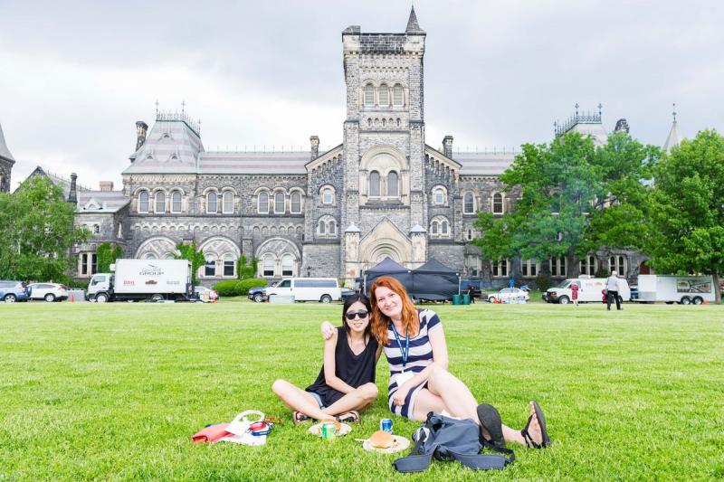 Two smiling women sit with a picnic lunch on the grass in front of University College.