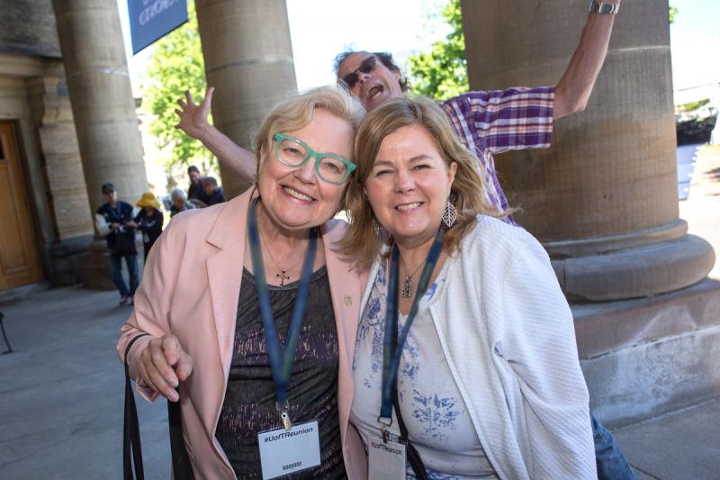 Two women pose for a picture with their arms around each other while a man behind them jumps up and makes a funny face.