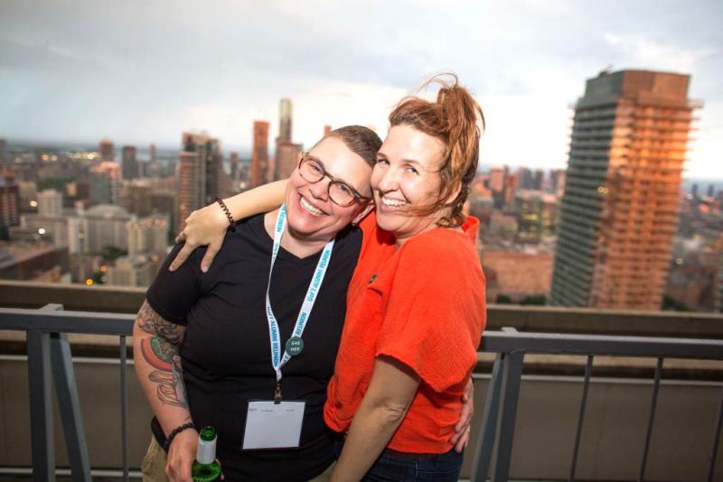 Two woman laugh with their arms around each other, standing on a balcony with a view out over Toronto.