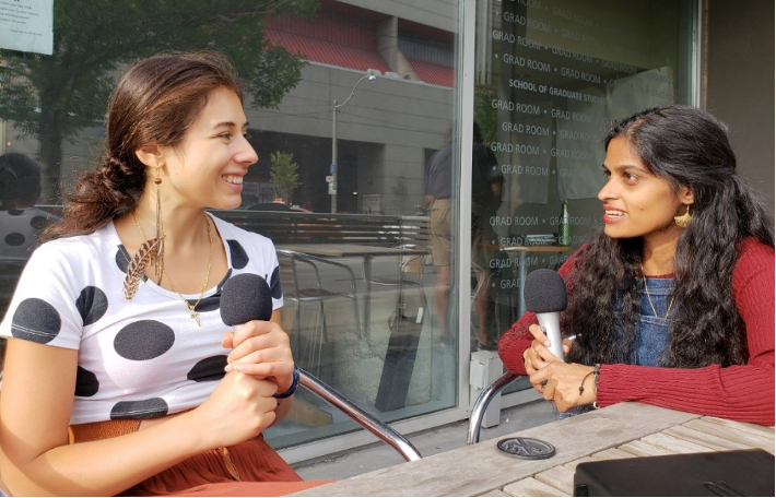 Swapna Mylabathula and Brenda Varriano sit at a café table outdoors, each holding a microphone, and chat together.