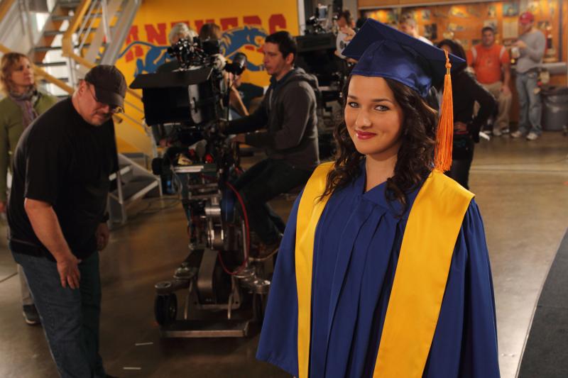 Annie Clark, in graduation robes, stands in front of a film camera and crew.