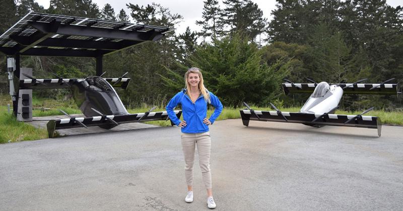Kristina Menton stands in front of two vehicles that look like race cars with no wheels and very long, wide, flat axles.