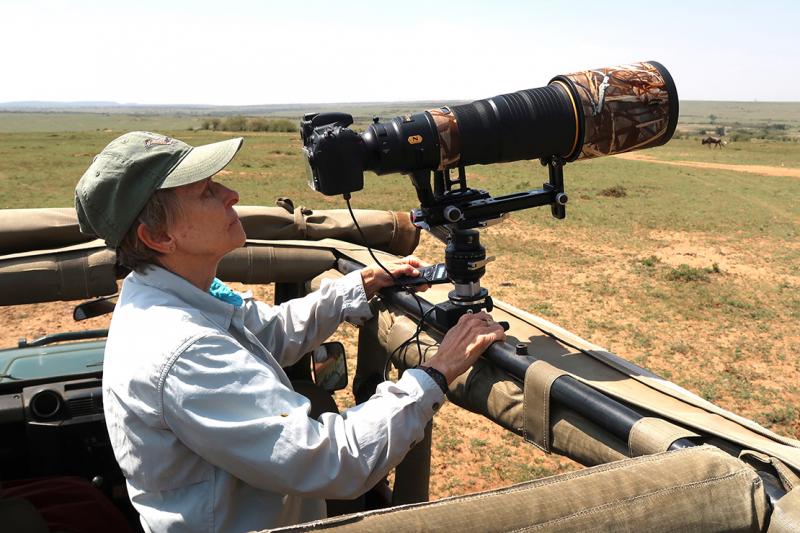 On an open plain, Roberta Bondar stands in the back of a jeep and looks through a large telephoto lens.