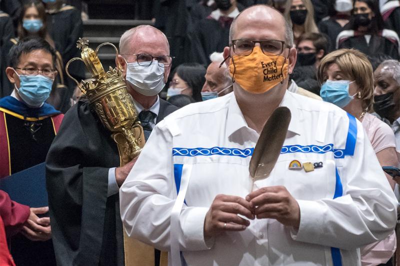 Jason Bazylak, wearing an every child matters mask, carries the eagle feather through Convocation Hall.