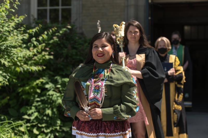 Tee Duke, wearing a ribbon skirt and beaded plaque, carries an eagle feather at the head of a convocation procession.