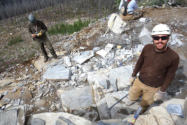 Cedric Aria smiles, wearing a hard hat and standing on rocky ground littered with slabs of flat stones.