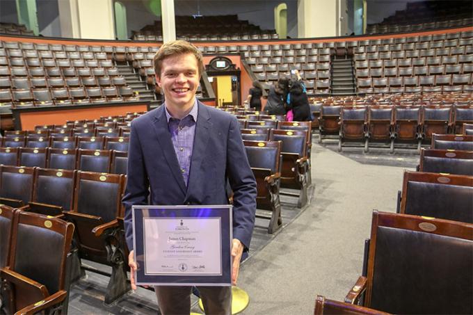 James Chapman stands in the middle of Convocation Hall, smiling and holding his Gordon Cressy Student Leadership Award