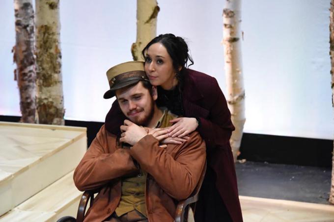 George Alevizos, wearing a 19th-century coat and army cap, holds the hand of a fellow actor standing behind him on stage.