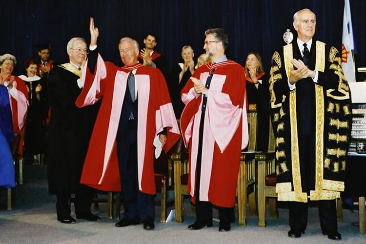 Christopher Plummer, wearing academic robes, waves from the stage at Convocation Hall as he stands next to Chancellor Hal Jackman.