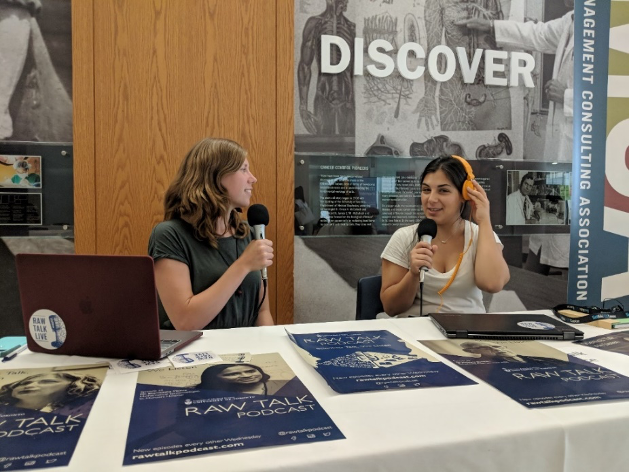 Grace Jacobs and Melissa Galati sit at a table covered with posters for the Raw Talk Podcast. Each holds a microphone.