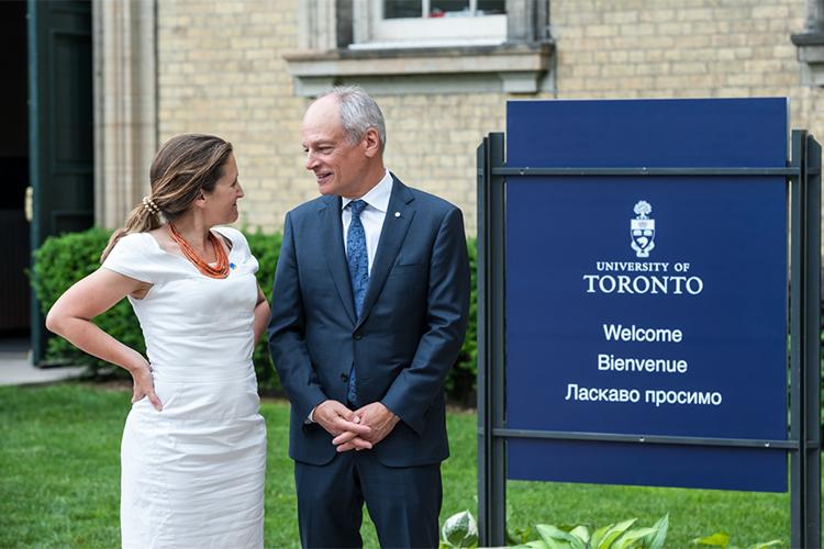 Chrystia Freeland Meric Gertler chat and look at each other while standing by a sign with the word "Welcome" in English, French and Ukrainian.