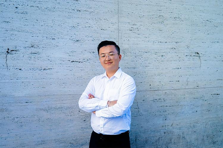 Richard Cao smiles, standing with arms folded in front of a stone wall.
