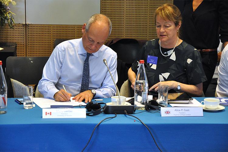 Meric Gertler signs a paper lying on a conference table as a delegate from the UK looks on.