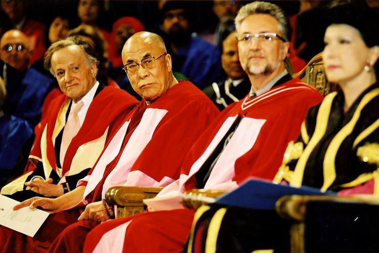 His Holiness the Dalai Lama wears academic robes over his monk's robe as he sits among dignitaries on the Convocation stage.