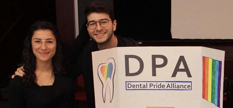 Lucia Santos and Evan Benak smile next to a poster with a rainbow-coloured tooth and the words: Dental Pride Alliance.