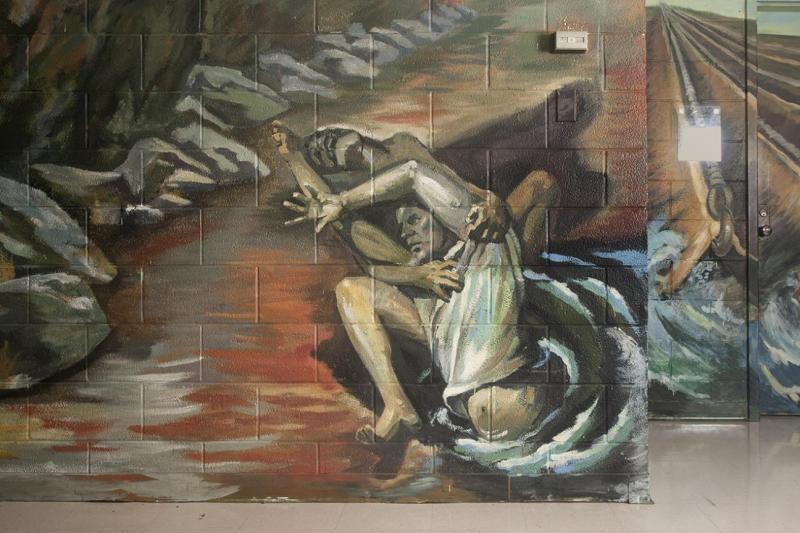 Painting on cinder block wall depicting a man reaching out from what appears to be water, with another behind holding him.