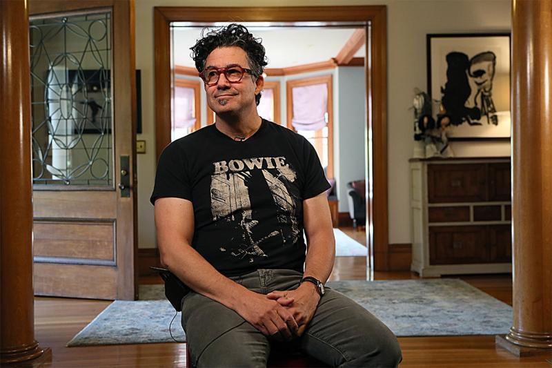 Derek Rossi, wearing a Bowie T-shirt and headphones, smiles as he sits in the hallway of a large house.