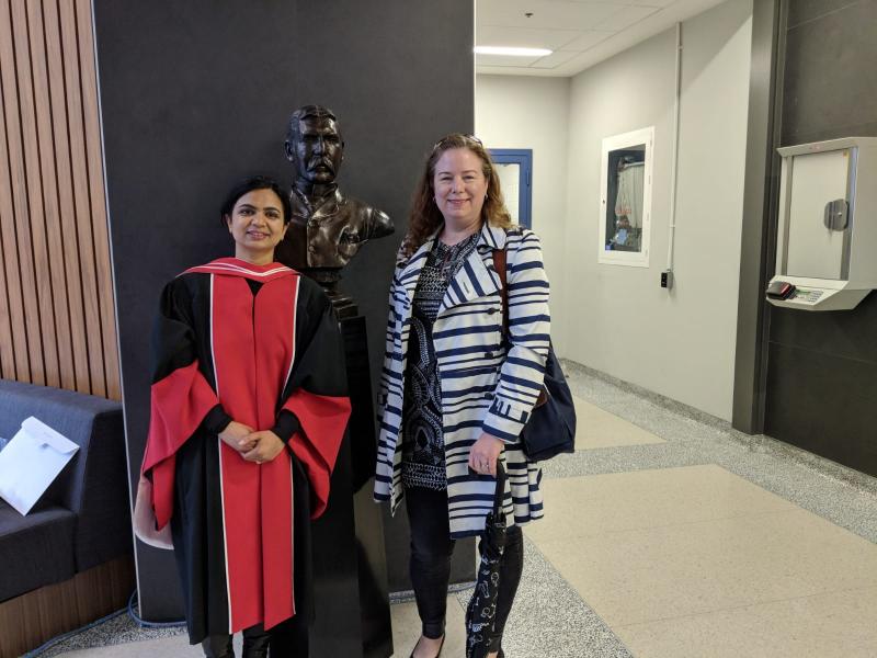 Navroop Dhaliwal, wearing academic robes for graduation, smiles with her professor Jennifer Mitchell in a hallway indoors.
